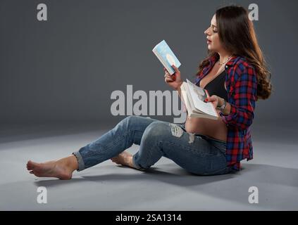 Eine schwangere Frau sitzt barfuß auf dem Boden und lächelt, während sie ein Buch über die Erziehung liest Stockfoto