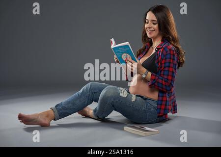 Eine schwangere Frau sitzt barfuß auf dem Boden und lächelt, während sie ein Buch über die Erziehung liest Stockfoto