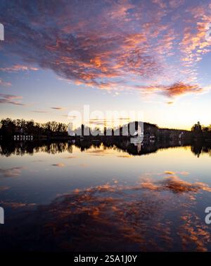 Ein ruhiger See bei Sonnenuntergang, der bunte Wolken und Bäume reflektiert. Ein kleines Gebäude ist am Ufer sichtbar, umgeben von Natur. Der Himmel ist mit gemalt Stockfoto