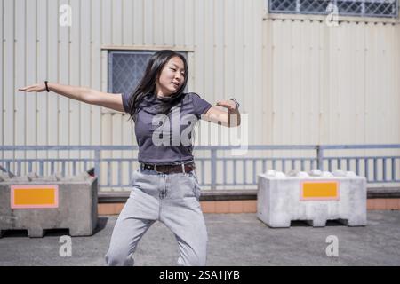 Weibliche junge asiatische K-Pop-Tänzerin, die sich energetisch in einem industriellen Stadtraum unter der Sonne bewegt Stockfoto