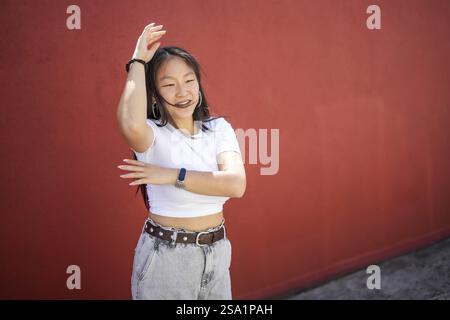 Glückliche energiegeladene asiatische Frau, die Arme hebt und einen urbanen Tanz im Freien vor einer roten Wand choreografiert Stockfoto