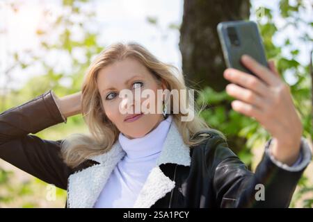Eine attraktive blonde Erwachsene Frau macht ein Selfie vor dem Hintergrund der Frühlingsnatur. Stockfoto