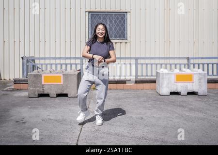 Frontales Foto einer lächelnden jungen asiatischen Frau, die K-Pop auf einem Industriedach tanzt Stockfoto