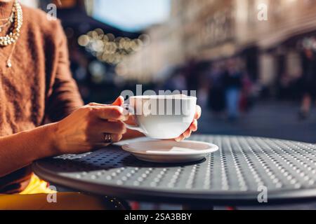 Nahaufnahme einer Tasse Kaffee in weiblichen Händen. Frau, die heißes Getränk trinkt, sitzt am Tisch im Café im Freien bei Sonnenuntergang. Leerraum Stockfoto