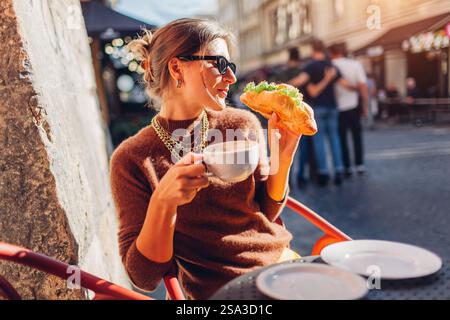 Junge Frau, die frisches Croissant und Kaffee im Café im Freien bei Sonnenuntergang trinkt. Der Gast genießt Streetfood auf der Terrasse in der Bäckerei Stockfoto