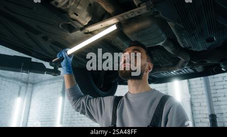 Kaukasischer Mann professioneller Mechaniker in Uniform Reparaturmann männlicher Ingenieur, der unter dem Auto arbeitet, überprüfen Sie das Problem unter dem Auto mit Taschenlampe beheben Stockfoto