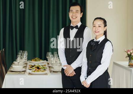 Porträt von Kellnern, die in die Kamera lächelten, während sie in einem eleganten Essbereich einen Dinner-Service einrichteten Stockfoto