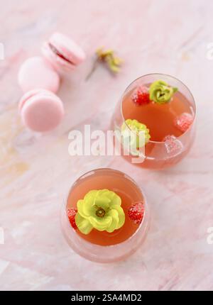 Zwei elegante Gläser halten ein erfrischendes Getränk mit Erdbeeren und essbaren Blumen, begleitet von zarten rosafarbenen Makronen auf einem glatten Marmor-sur Stockfoto
