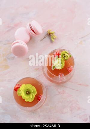 Zwei leuchtende Mocktails, die mit Erdbeeren und essbaren Blumen geschmückt sind, stehen neben zarten rosa Makronen vor einer ruhigen, hellrosa Marmorkulisse. Stockfoto