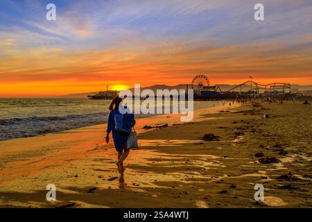 Frau, die bei Sonnenuntergang barfuß am Strand spaziert und den farbenfrohen Himmel und die Silhouette des Santa Monica Piers mit seinem Riesenrad und seiner Roller genießt Stockfoto