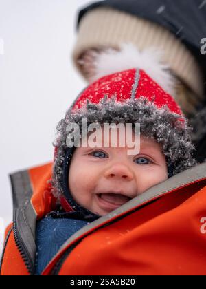 Glückliches Baby genießt den Schneetag im gemütlichen roten Hut während des Winterausflugs. Stockfoto