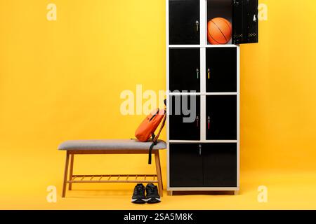 Offenes Schließfach mit Basketballball, Bank, Rucksack und Sneakers auf orangefarbenem Hintergrund Stockfoto