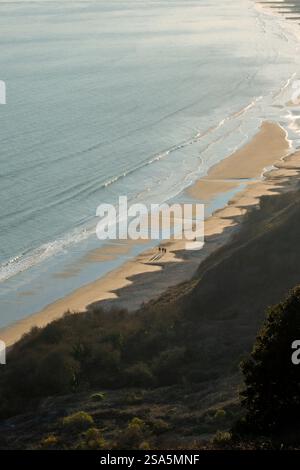Drei Personen genießen einen friedlichen Spaziergang entlang des Swanage Beach in Purbeck, während die Sonne untergeht und lange Schatten auf den goldenen Sand wirft Stockfoto
