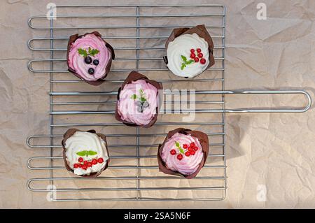 Köstliche Cupcakes mit cremigem Frosting und Beeren, die auf einem Gitter gekühlt werden. Frisch gebackene Leckereien mit rosa und weißer Glasur. Stockfoto