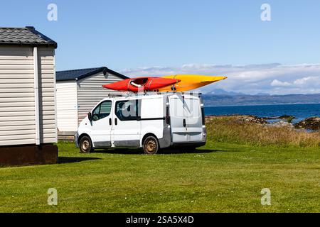Der alte Quirl eines weißen Lieferwagens, der an einem sonnigen Tag auf dem Gras geparkt war, mit zwei Kanus, die an den Dachträger geschnallt waren. Seehügel, Berge im Hintergrund Stockfoto