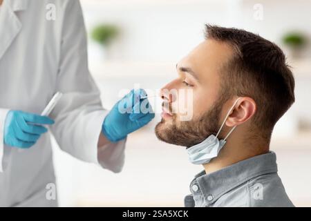 Covid-Diagnoseverfahren. Arzt, der nasalen PCR-Test für jungen kaukasischen Mann mit sterilen Abstrichstäbchen zu Hause macht Stockfoto