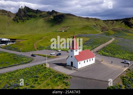 Ein malerischer Blick auf die isländische Kirche in Vik in der Nähe des schwarzen Strandes reynisfjara, umgeben von grünen Hügeln und lebendigen Wildblumen in Island Stockfoto