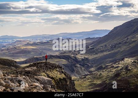 Einsamer Wanderer in roter Jacke auf einer felsigen Klippe mit Blick auf die weite Berglandschaft Islands Stockfoto
