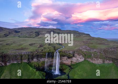 Aus der Vogelperspektive des majestätischen Seljalandsfoss Wasserfalls, der durch grüne Klippen unter leuchtenden Wolken bei Sonnenuntergang im Bereich des Golden Circle in Island kaskadiert Stockfoto