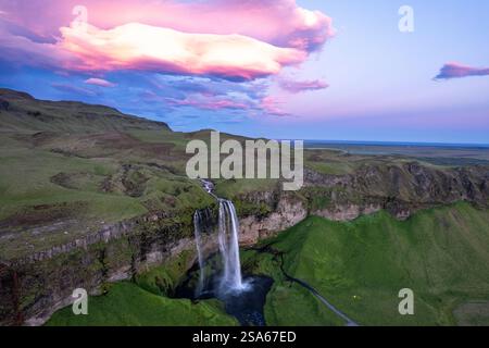Aus der Vogelperspektive des majestätischen Seljalandsfoss Wasserfalls, der durch grüne Klippen unter leuchtenden Wolken bei Sonnenuntergang im Bereich des Golden Circle in Island kaskadiert Stockfoto
