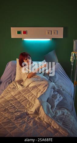 Junge lächelnde rothaarige Frau, die im Krankenhausbett liegt, Laptop hält und Online-Videoanrufe mit Familie und Freunden hat. Wiederherstellung und Support Stockfoto