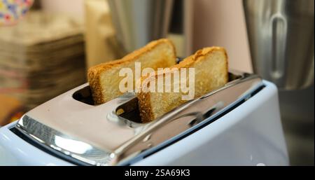 Goldenes geröstetes Brot wird aus einem blauen Toaster mit knuspriger Textur und warmer Ausstrahlung zubereitet. Der Toast ist frisch zubereitet für das Frühstück oder den Snack am Morgen Stockfoto