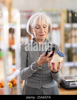 Ältere Käuferinnen scannen QR-Code auf Flaschenmayonnaise-Etikett in der Lebensmittelabteilung des Supermarktes Stockfoto