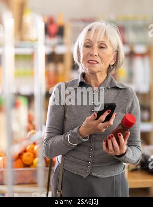 Ältere Käuferinnen scannen QR-Code auf Flaschenketchup-Etiketten in der Lebensmittelabteilung des Supermarktes Stockfoto