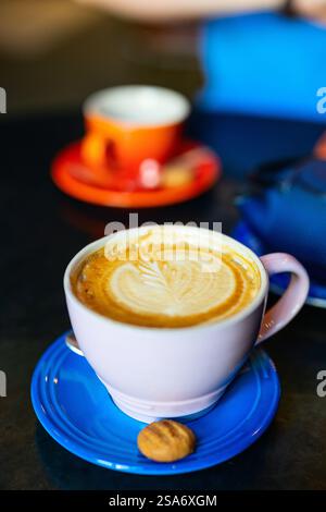 Weiße Kaffeetasse mit Latte mit herzförmiger Milchschaumkunst, serviert auf einer hellblauen Untertasse mit kleinem Keks. Stockfoto