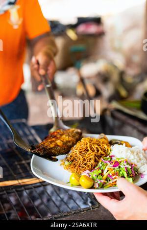 Eine überdachte Mahlzeit mit gegrilltem Fisch, gebratenen Nudeln, gedämpftem Reis und frischem Gemüsesalat mit Zitrusgarnitur, die auf einem Grill serviert wird. Stockfoto