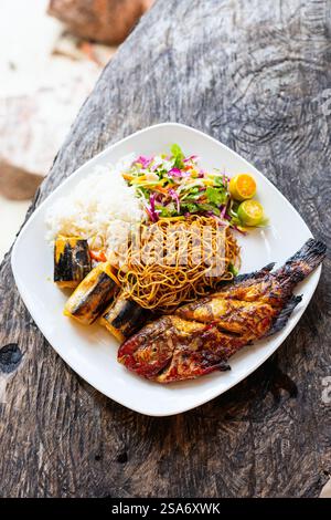 Ein köstlicher Teller mit gegrilltem Fisch, gebratenen Nudeln, gedämpftem Reis, frischem Salat, gegrillten Bananen, und Zitrusgarnitur, serviert auf einem rustikalen Wald Stockfoto