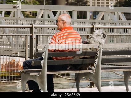 Ein älterer Mann sitzt mit seinem Hund auf einer Bank auf einem Dock im Stadtteil Bay Ridge in Brooklyn, Brooklyn, New York, USA, 20. September 2023 Stockfoto
