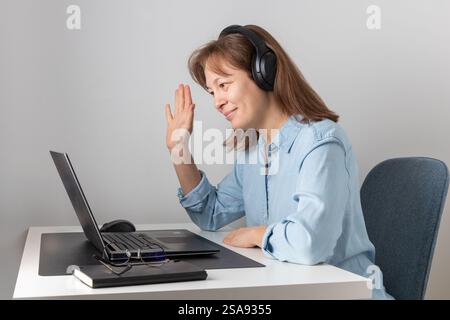 Fröhliche freiberufliche Frau in großen Kopfhörern, die bei Videokonferenzgesprächen spricht, mit der Hand zum Laptop winkt, Hallo sagt, lächelt, lacht Stockfoto
