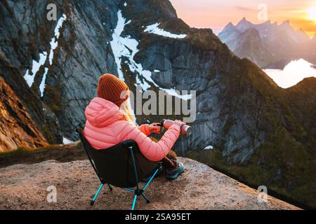 Frau, die in Norwegen reist und sich im Campingstuhl mit Thermoskanne entspannt, Wandern im Freien, Mädchen trinkt heißen Kaffee genießt Sonnenuntergang Lofoten Inseln auf Reinebrin Stockfoto