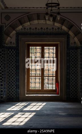 Reich verzierte Fenster mit komplizierten Holzgittern und traditionellen osmanischen Fliesen in einer historischen Moschee. Sonnenlicht wirft geometrische Schatten auf den Boden Stockfoto
