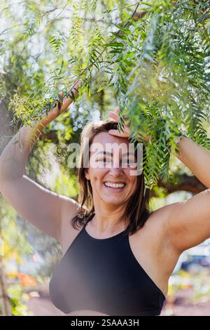 Eine Frau lächelt und posiert vor einem Baum. Sie trägt ein schwarzes Oberteil und genießt die Natur Stockfoto