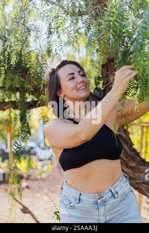 Eine glückliche Frau steht unter einem Baum, berührt die Blätter und lächelt. Stockfoto