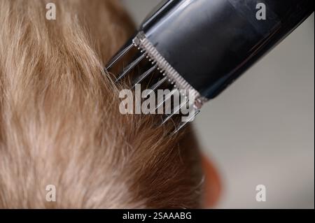 Haarschnitte aus blondem Haar für einen Jungen mit einem Haarschneider, Nahaufnahme. Haarpflegekonzept. Mutter schneidet dem Sohn zu Hause mit einem Haarschneider die Haare. Stockfoto