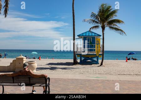 Mann, der sich auf einer Bank am Strand mit einem Rettungsschirm und Palmen in Hollywood Beach, Florida, entspannt. Stockfoto