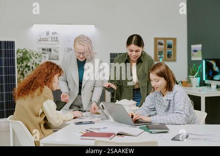 Gruppe verschiedener Kollegen, die an einem modernen Bürotisch zusammenarbeiten und Projektdetails diskutieren, wobei jeder mit Laptops und Tablets interagiert und Ideen erarbeitet Stockfoto