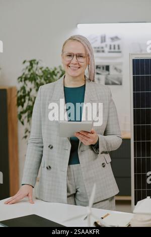 Porträt einer lächelnden Frau mit Brille und Tablette im modernen Büro. Geschäftsumfeld mit professionellem Setting und Hintergrundelementen einschließlich Pflanzen Stockfoto