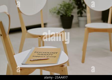 Gedämpfte Therapieeinrichtung mit mehreren leeren Holzstühlen, die im Kreis um einen kleinen Tisch angeordnet sind, der das Psychologietherapiezeichen in angenehmer Umgebung hält Stockfoto