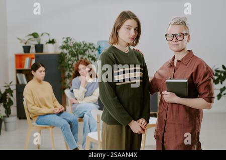 Gruppe von Einzelpersonen, die an einer Beratungssitzung in einem gut beleuchteten modernen Büro teilnehmen. Zwei Frauen stehen, während andere Teilnehmer im Hintergrund sitzen und diskutieren Stockfoto