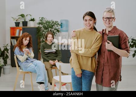 Gruppe aus vier verschiedenen Kollegen, die sich in einem gemütlichen Bürobereich entspannen und Zuneigung und Gesellschaft zeigen, mit Bücherregalen und Büromaterialien im Hintergrund Stockfoto