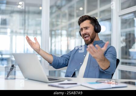 Unzufriedener Mann sitzt im Büro an seinem Arbeitsplatz in Kopfhörern, Mitarbeiter schaut unzufrieden auf den Laptop-Bildschirm, sieht erfolglose Sportspiele an, jubelt. Stockfoto