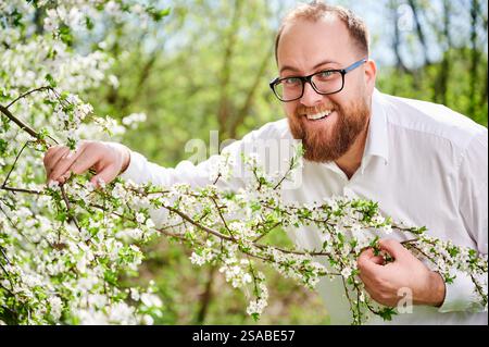 Mann-Allergie genießt nach Behandlung von Saisonallergie im Frühjahr. Porträt eines glücklichen bärtigen Mannes, der im Frühling vor einem Blütenbaum lächelt. Frühjahrsallergiekonzept. Stockfoto