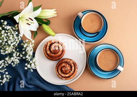 Zimtrollen auf einem weißen Teller mit zwei Tassen Kaffee in blauen Keramikbechern, weißen Lilien und gypsophila-Blüten auf einem beigen Hintergrund mit Stockfoto