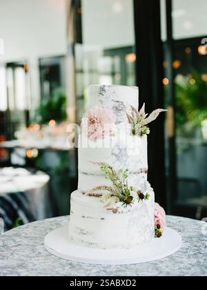 Eine atemberaubende dreistufige Hochzeitstorte mit rustikalem Frostdesign mit zarten Blumen und Grün. Die elegante Umgebung schafft ein romantisches Ambiente Stockfoto