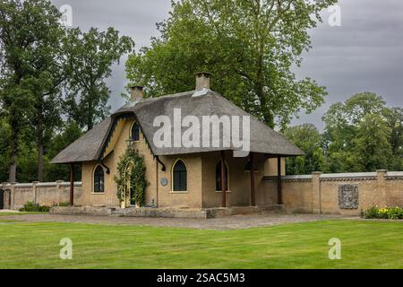 Ein charmantes Backsteinhaus mit einem Strohdach, bogenförmigen Fenstern und einer grünen Tür, die mit Kletterpflanzen bedeckt ist. Das Haus liegt in einem üppigen Garten mit einem cobb Stockfoto