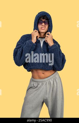 Modischer Mann mit blauem, flauschigem Hoodie, Sonnenbrille und Jogginghose, posiert auf gelbem Hintergrund Stockfoto
